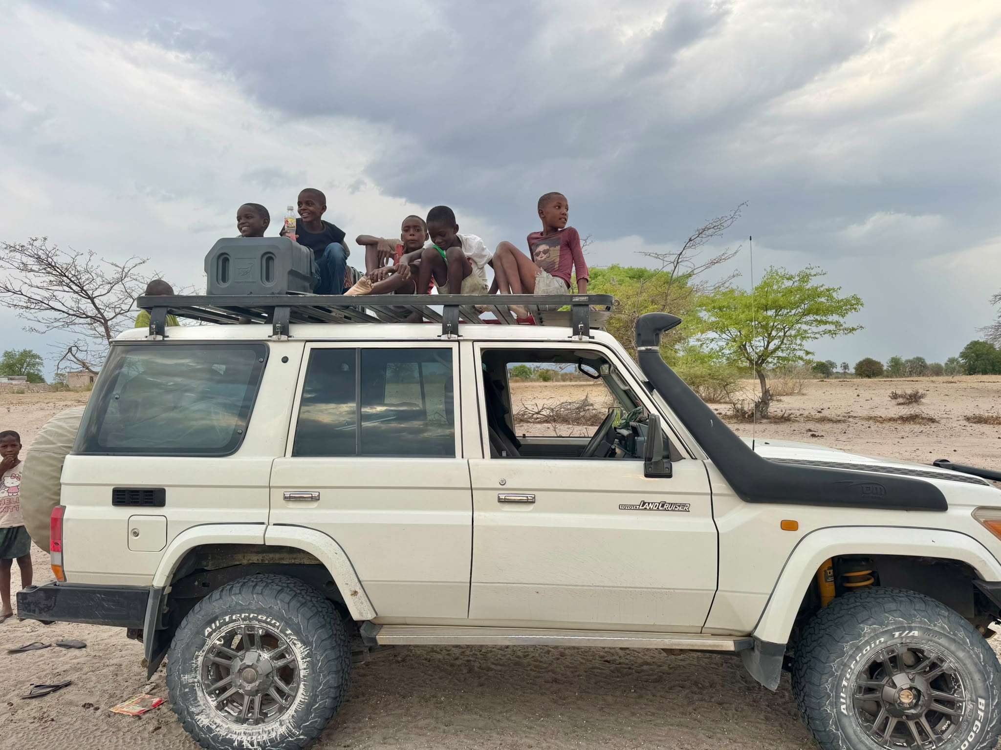 Children sitting on the roof rack of a white Land Cruiser beneath a stormy sky